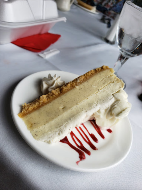 A slice of vanilla cheesecake on a plate, topped with whipped cream and drizzled with raspberry sauce.