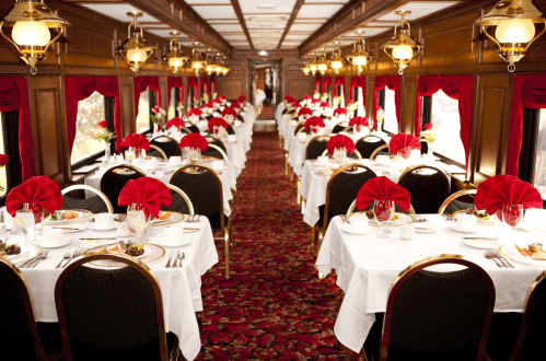Elegant dining car with tables set for a formal meal, featuring white tablecloths, red napkins, and floral centerpieces.