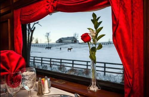 A dining table with a rose in a vase, overlooking a snowy landscape with horses and a farmhouse through a window.