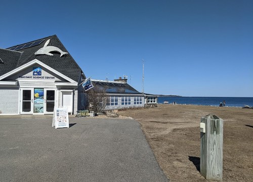 A coastal science center building near the beach, with a clear blue sky and ocean in the background.