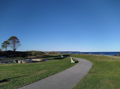 A winding path through a grassy area leads to the ocean under a clear blue sky. Benches are visible along the path.