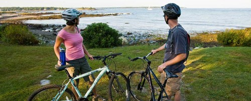 A man and woman in helmets stand with bicycles, looking at a scenic view of the ocean and shoreline.
