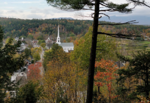 A scenic view of a small town with a church steeple, surrounded by autumn foliage and distant mountains.