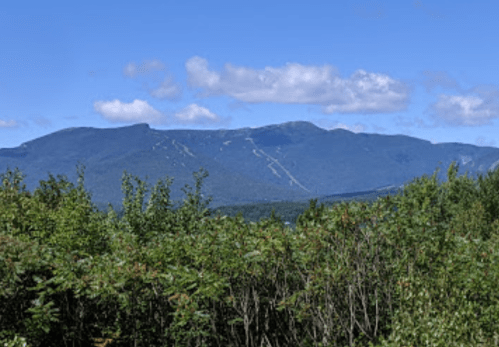 A scenic view of a mountain range under a blue sky, surrounded by lush greenery and scattered clouds.