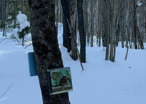 A snowy forest scene with a tree displaying a colorful animal painting and a blue sign in the background.