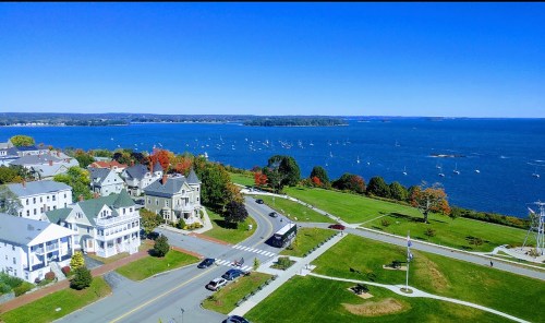 A scenic view of a coastal town with colorful trees, a blue bay, and sailboats on a clear day.