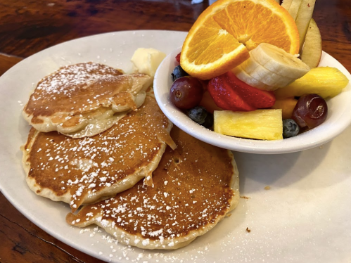 A plate of fluffy pancakes dusted with powdered sugar, served with a bowl of fresh fruit including orange, banana, and berries.