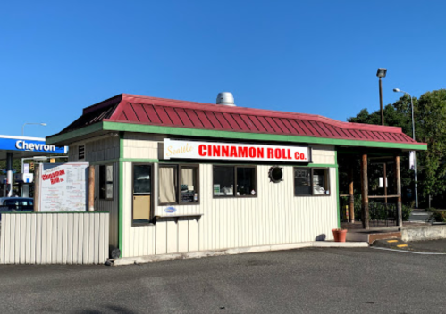 A small, colorful building with a red roof, featuring a sign that reads "Seattle Cinnamon Roll Co." in bright letters.
