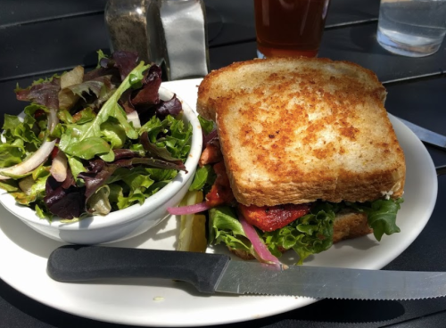 A plate with a crispy sandwich, pickles, and a side salad of mixed greens on a table.