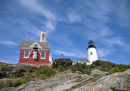 A red and white lighthouse beside a rocky shore under a blue sky with scattered clouds.