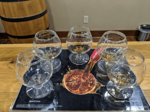 Five glasses on a wooden bar top, with one being filled with a golden liquid, and a Maker's Mark mat underneath.
