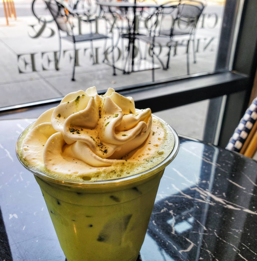 A close-up of a matcha drink topped with whipped cream, sitting on a marble table with a café window in the background.