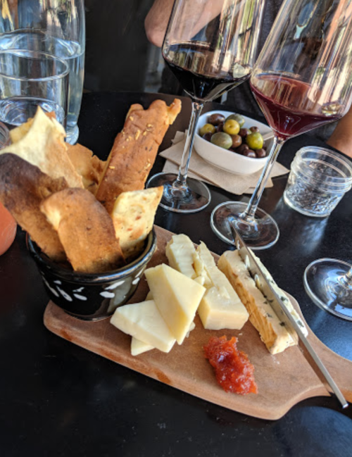 A wooden board with assorted cheeses, a bowl of crackers, olives, and two glasses of red wine on a table.
