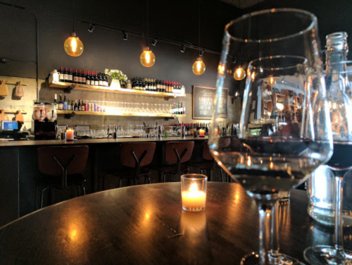 A cozy bar scene with wine glasses, a candle, and shelves lined with bottles, illuminated by warm pendant lights.