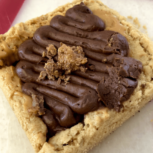 A close-up of a dessert bar topped with chocolate frosting and crumbled toppings on a light-colored base.