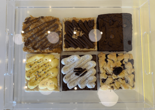 A display of six assorted dessert bars, including caramel, chocolate, and cookie flavors, in a clear container.