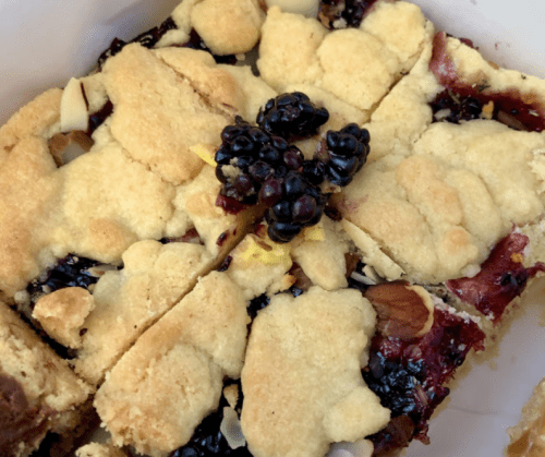 A close-up of a baked dessert with a crumbly top, featuring blackberries and a berry filling, cut into squares.
