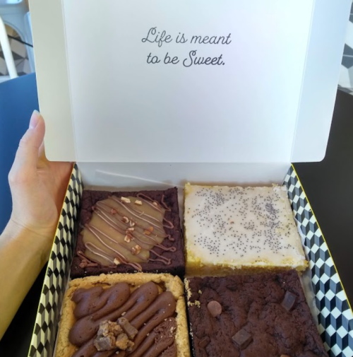 A hand holding a box of assorted brownies and dessert bars with the text "Life is meant to be Sweet" inside the lid.