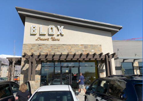 Exterior of Blox Dessert Bar with a stone facade and large windows, featuring a clear blue sky and parked cars.