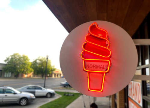 Neon sign of a soft serve ice cream cone with the word "NORMAL" in the center, glowing against a bright sky.