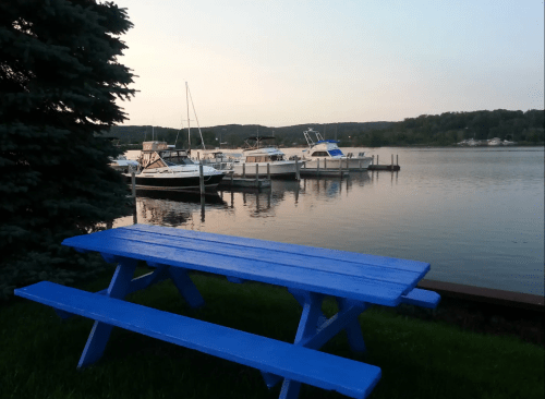 A blue picnic table by a calm lake, with boats docked in the background and trees lining the shore.
