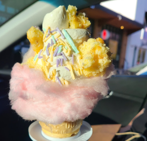 A colorful ice cream cone topped with cotton candy, sprinkles, and chunks of cake, set against a blurred background.
