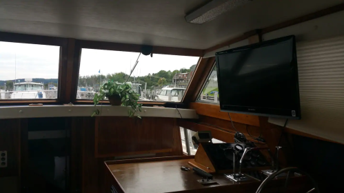 Interior of a boat with a wooden table, a TV mounted on the wall, and a view of the marina through large windows.