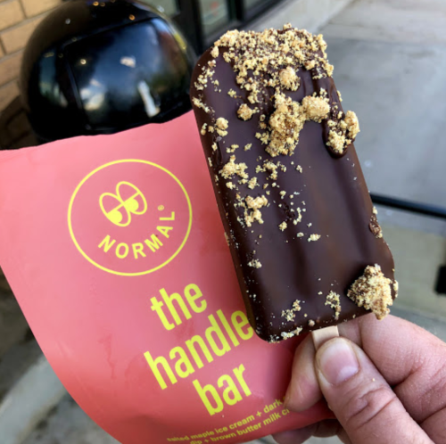 A hand holds a chocolate-covered ice cream bar topped with crumbs, next to a pink package labeled "the handle bar."