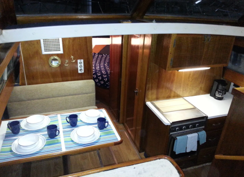 Interior of a boat featuring a dining area with plates and mugs, a kitchen counter, and a doorway to a sleeping area.