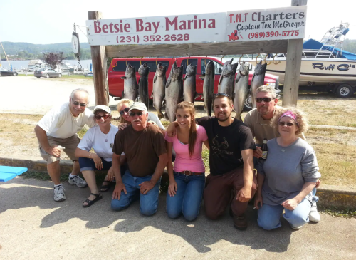 A group of eight people poses in front of a sign at Betsie Bay Marina, showcasing their catch of fish hanging on display.