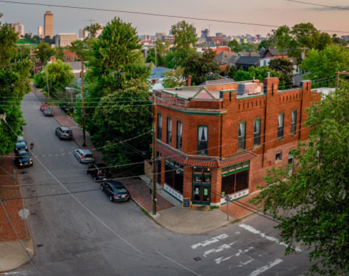 A brick building at a street corner, surrounded by trees and parked cars, with a city skyline in the background.