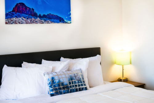 A cozy bedroom featuring a bed with white linens, a decorative pillow, and a lamp beside a mountain landscape artwork.