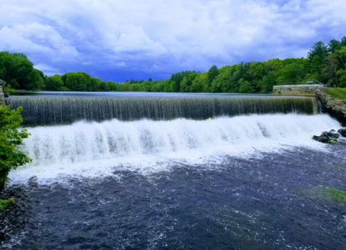A serene waterfall cascades over a dam, surrounded by lush green trees under a cloudy sky.