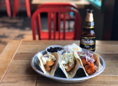 Three tacos filled with fish, topped with cabbage and onions, served with rice, black beans, and a bottle of Modelo beer.