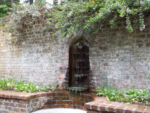 A small wall fountain with water cascading down, surrounded by greenery and brick landscaping.