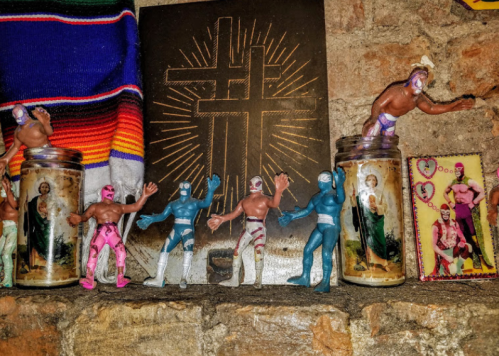 Colorful lucha libre figurines and candles with religious images displayed on a rustic brick wall.