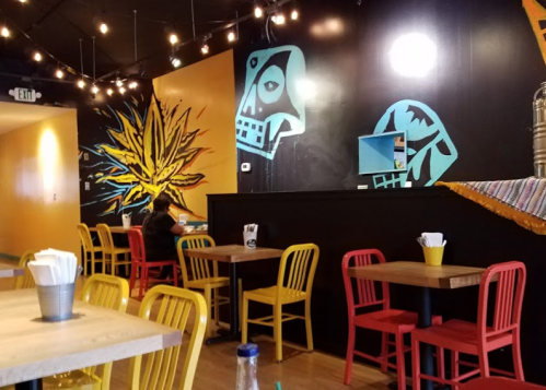 Colorful restaurant interior featuring vibrant wall art, wooden tables, and colorful chairs. A person sits at a table.