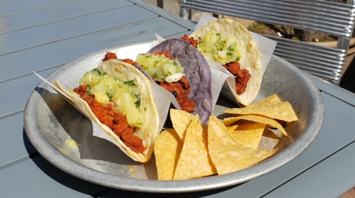 Three tacos with meat and pineapple on a metal plate, accompanied by tortilla chips.