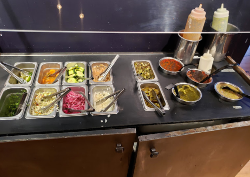 A variety of condiments and toppings in metal trays, including vegetables and sauces, on a dark countertop.