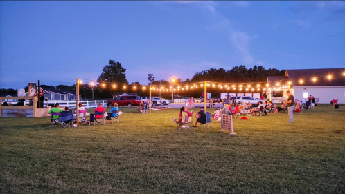 A lively outdoor gathering at dusk, with string lights illuminating people sitting in chairs on a grassy field.