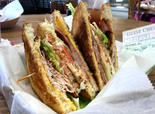 A stacked sandwich with layers of turkey, ham, lettuce, and tomato, served on toasted bread with a guest check beside it.