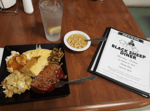 A plate of meatloaf, mashed potatoes, green beans, macaroni, cornbread, and a side of beans, with a drink and menu.