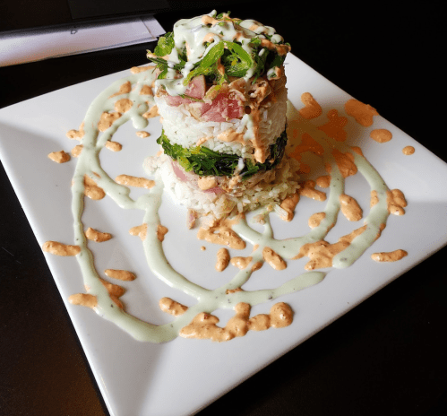 A layered sushi dish on a square plate, featuring rice, greens, and sauces drizzled around it.