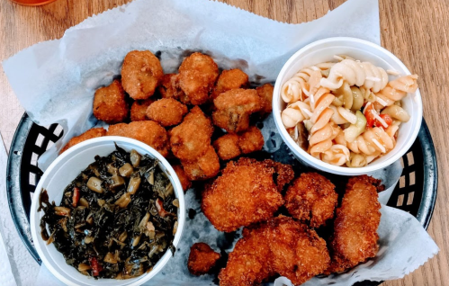 A basket with fried food, including crispy chicken, collard greens, and pasta salad in separate cups.