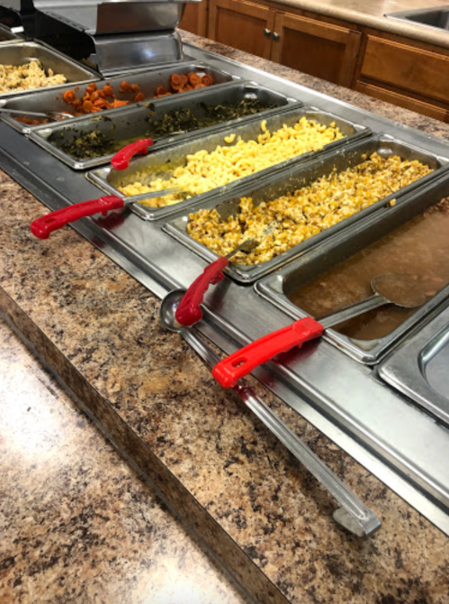 A buffet with various food trays, including macaroni, greens, and carrots, featuring red serving utensils.