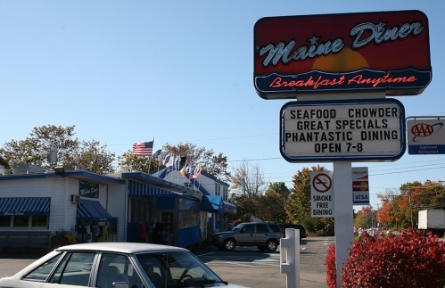 A diner with a sign advertising seafood chowder and specials, surrounded by trees and flags, with a car parked outside.