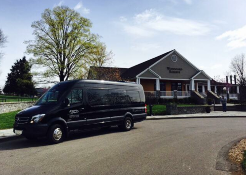 A black van parked in front of a building with a large porch and trees on a sunny day.