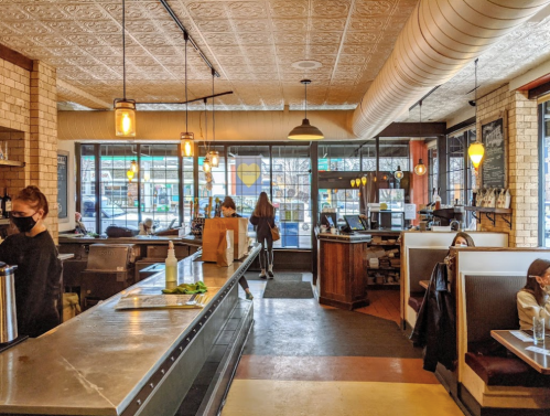 A cozy restaurant interior with wooden accents, booths, and patrons, featuring large windows and warm lighting.