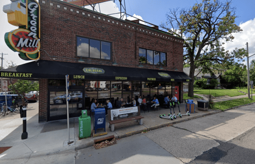 A bustling café with outdoor seating, featuring a brick exterior and large windows, surrounded by trees and scooters.