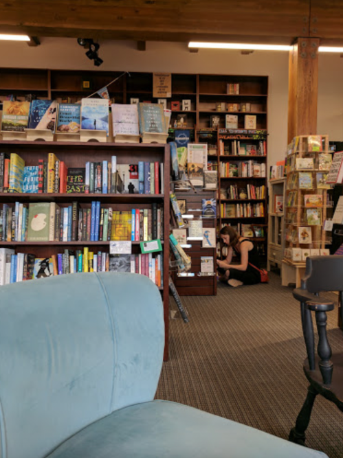 A cozy bookstore interior with bookshelves, a blue chair in the foreground, and a person browsing on the floor.